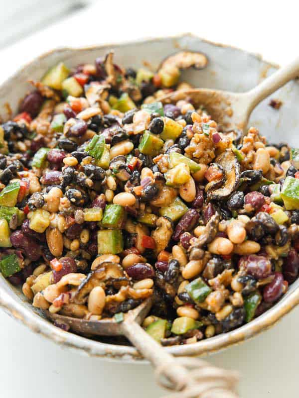 A bowl filled with a colorful dense bean salad containing black beans, kidney beans, navy beans, chopped cucumber, diced red pepper, and mushrooms, mixed together with a Korean BBQ dressing. A spoon rests in the bowl.